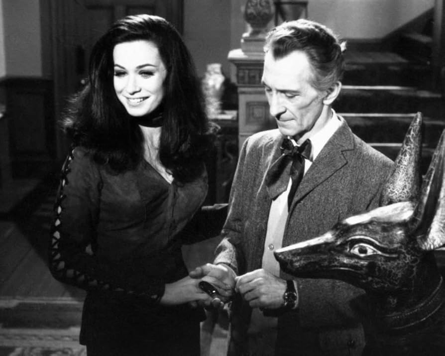 Foto em preto e branco de bastidores de Valerie Leon (sorridente) contracenando com Peter Cushing, que segura sua mão ao colocar uma joia em seu dedo.