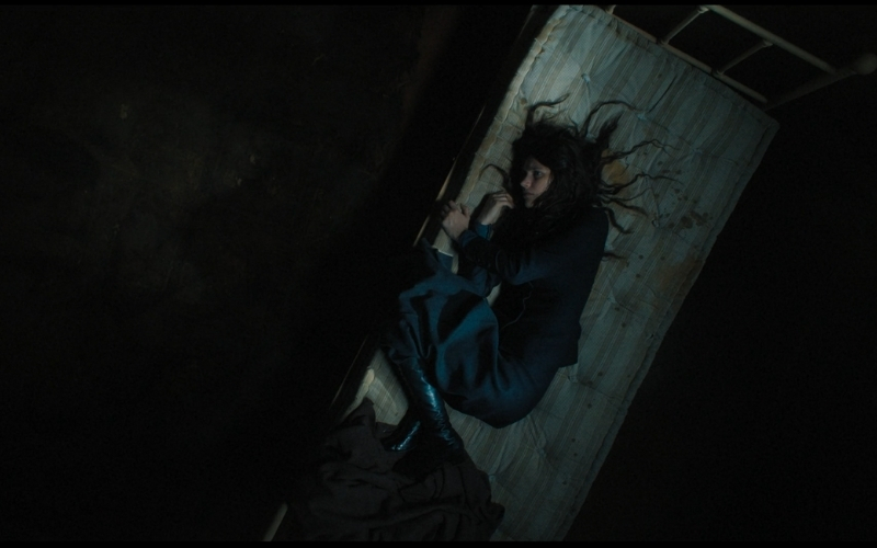 Cena do filme A Primeira Profecia (2024) mostrando Margaret deitada em um pequeno colchão em posição fetal no quarto do castigo. Seus cabelos novamente estão revoltos.