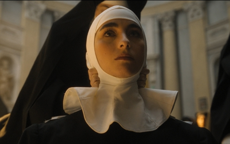 Cena do filme A Primeira Profecia (2024) mostrando a personagem Luz em sua cerimônia para tornar-se freita. Atrás dela, uma mulher aperta a coifa em sua cabeça, enquanto Luz está com uma expressão séria.