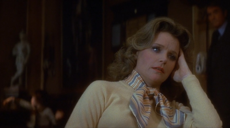 Cena do filme A Profecia (1976) mostrando a personagem Katherine em primeiro plano, sentada e com sua cabeça apoiada na sua mão. Ela possui um olhar vago. Atrás dela, em segundo plano, aparece a criança anticristo Damien brincando.