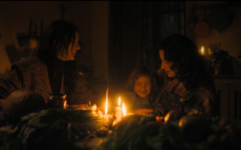 Cena do filme A Primeira Profecia (2024) mostrando Margaret, sua filha e Carlita rindo e jantando reunidas em uma casa de madeira.