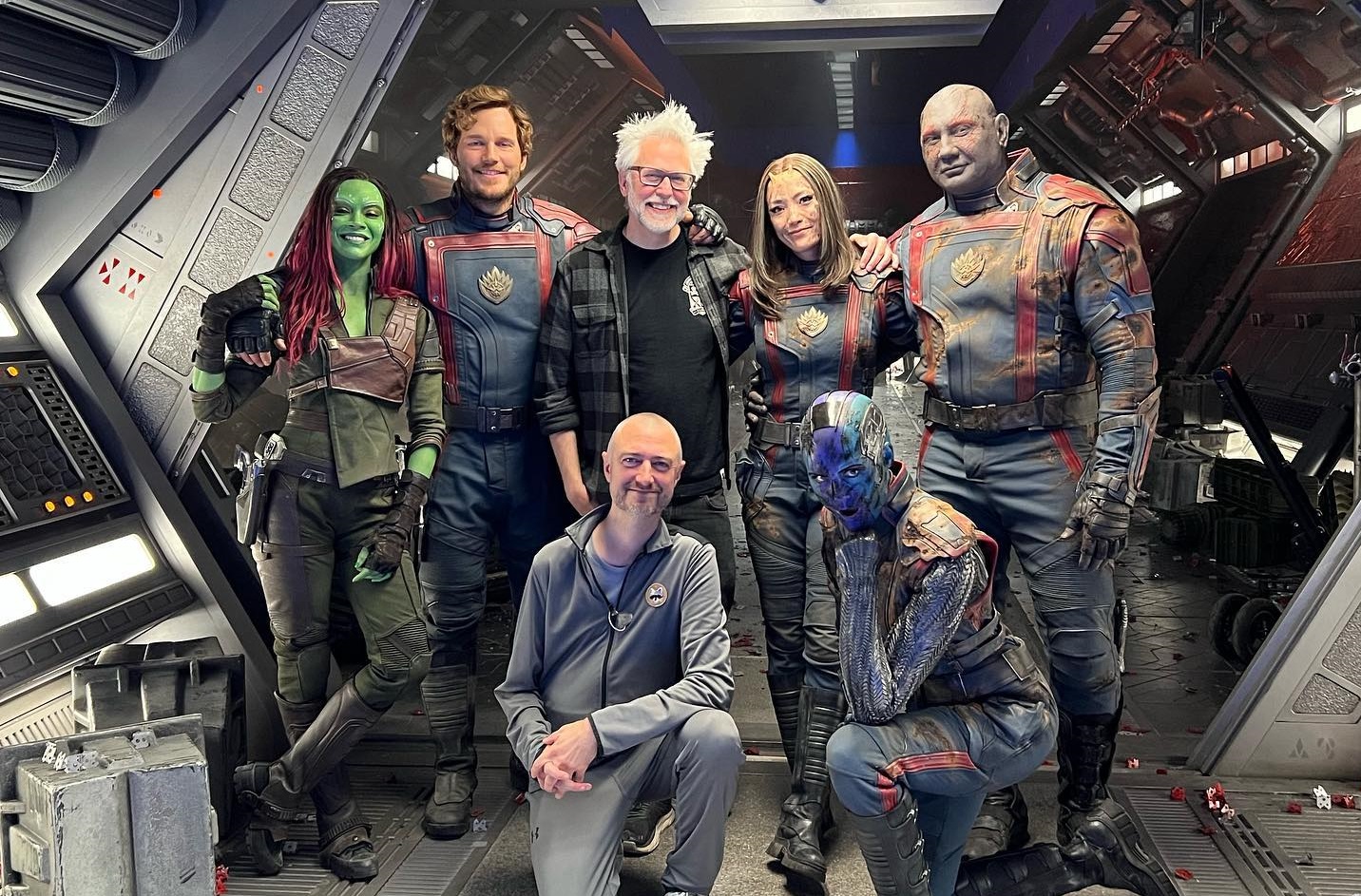 Foto do set de Guardiões da Galáxia Vol. 3 mostrando os atores caracterizados ao lado de James Gunn. Da esquerda para a direita: Gamora (Zoe Saldaña), Senhor das Estrelas (Chris Pratt), James Gunn, Sean Gunn, Mantis (Pom Klementieff), Nebulosa (Karen Gillan) e Drax ( Dave Bautista).