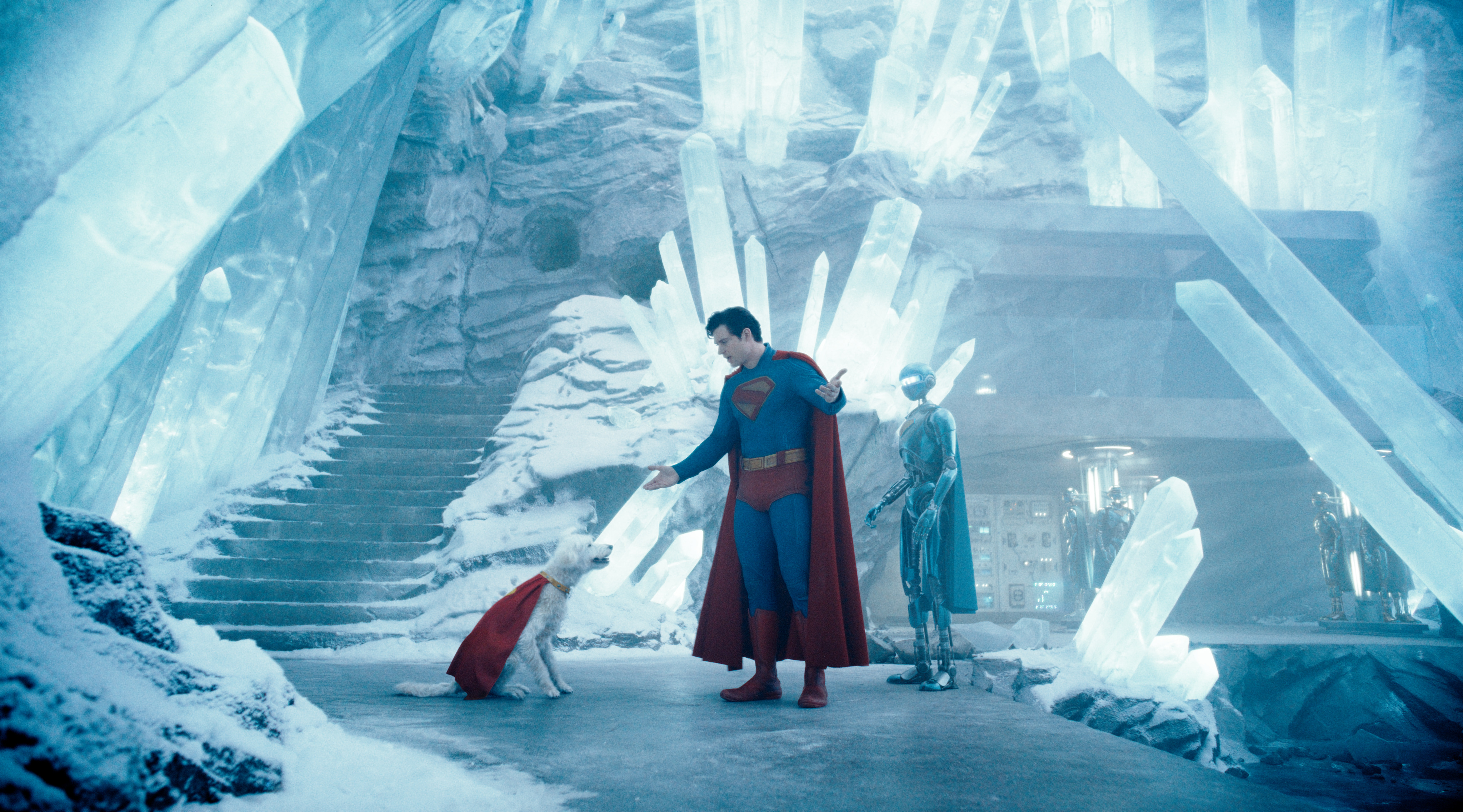 Cena do filme Superman (2025) mostrando o Superman de pé, discutindo com o cachorro Krypto que está sentado o ouvindo. Ao seu lado há um robô e todos estão em meio aos cristais da morada do Superman, a Fortaleza da Solidão, toda azul e reluzente.