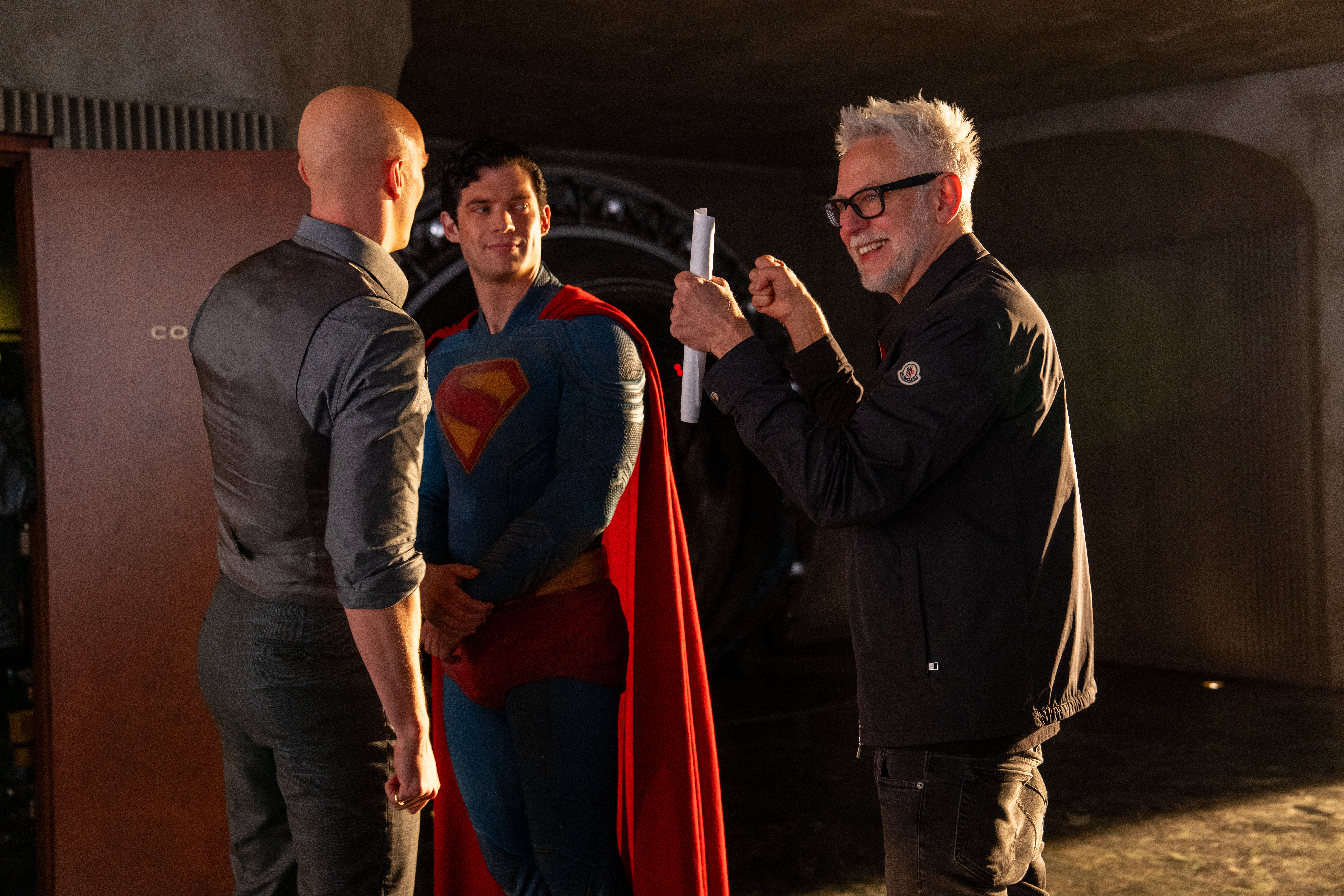 Foto do set de Superman (2025) mostrando Lex Luthor (Nicholas Hoult) careca e de roupa social de costas, no meio está Superman (David Corenswet) trajado com uniforme e capa, olhando para James Gunn que está de pé e sorridente ao lado deles gesticulando com as mãos.
