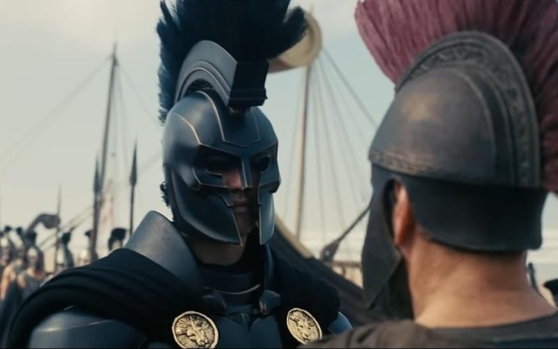 Cena do filme A Odisseia. Mostra Agamemnon encarando Odisseu, ele veste uma armadura negra, polida e se destaca dos outros soldados gregos.