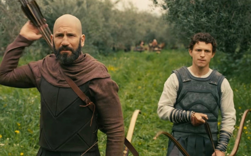 Cena do filme A Odisseia. Menelau e Telêmaco estão em um campo verde segurando arcos e flechas. Menelau parece vestir um moletom bordô com um colete de borracha