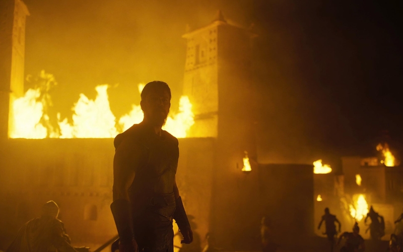 Cena do filme A Odisseia. Odisseu está olhando para o incêndio em Troia à noite. Há torres e casas pegando fogo.