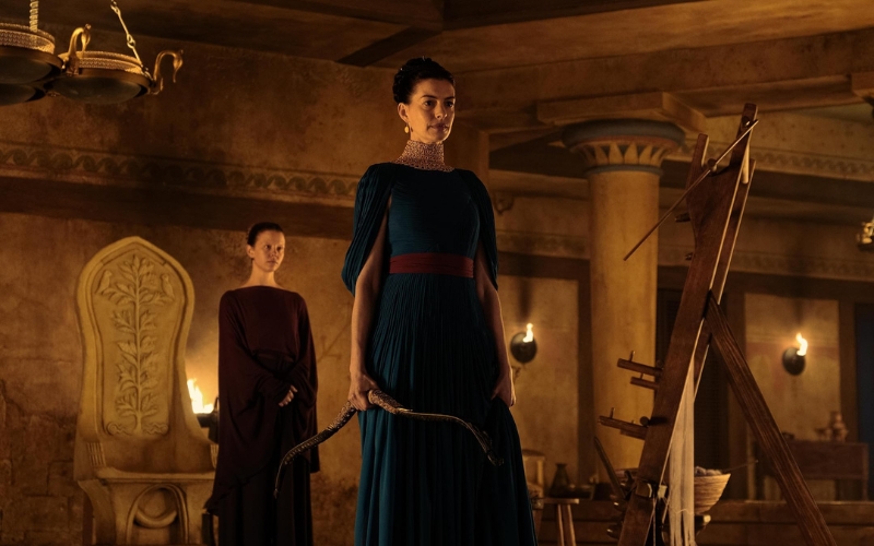 Cena do filme A Odisseia. Penélope e Melanto no banquete em Ítaca. Penélope está em pé segurano o arco de Odisseu.