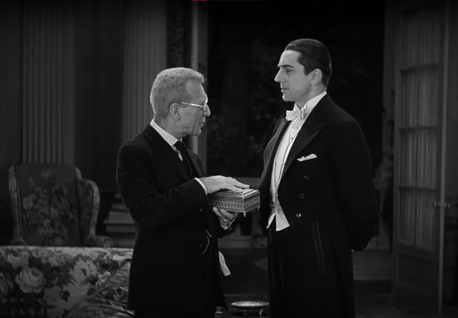 Cena do Filme Drácula de 1931 com Van Helsing, velho e de óculos, segurando uma caixa em frente à Drácula, que o encara com expressão altiva.