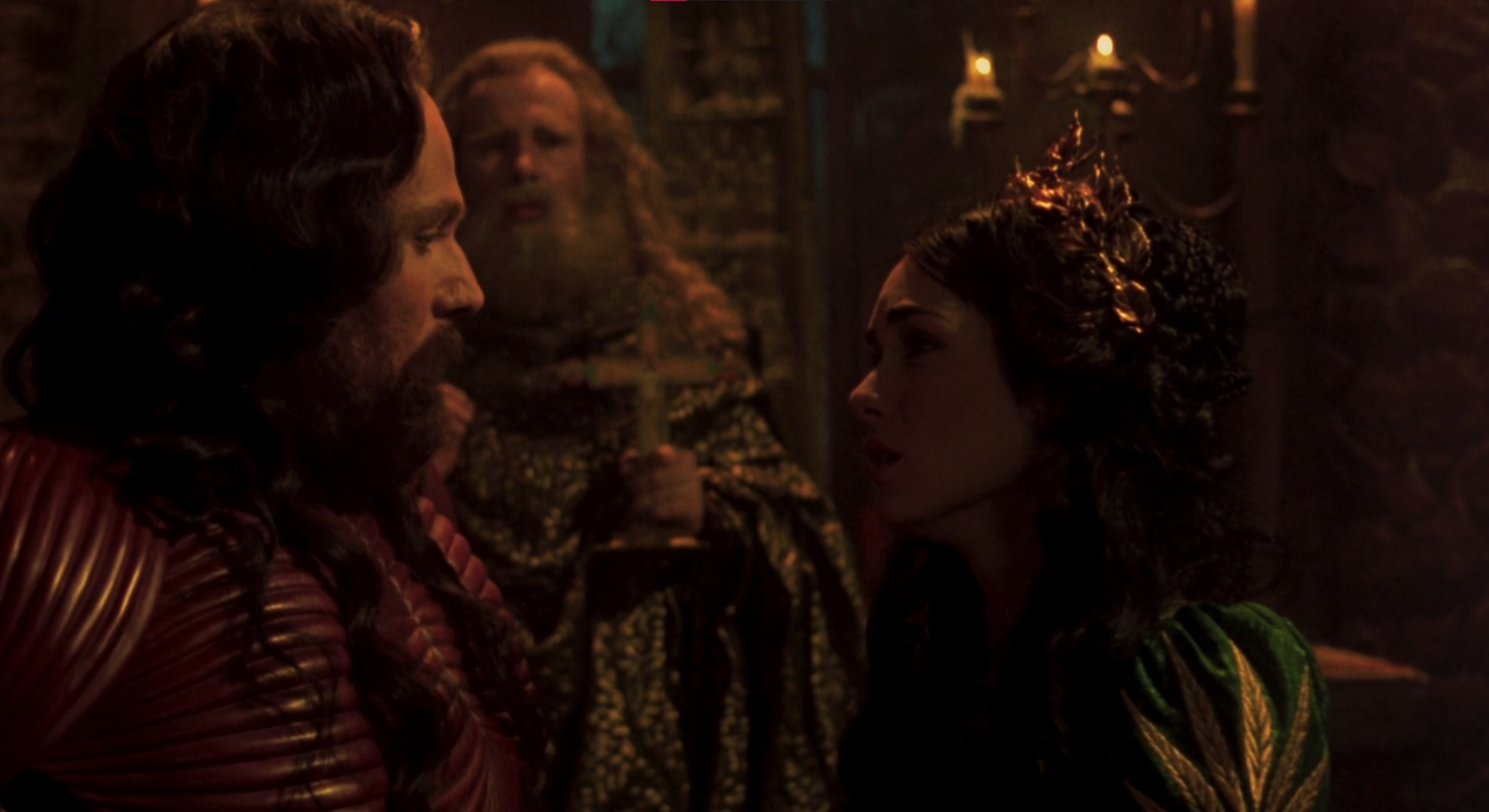 Cena do Filme Drácula de Bram Stoker mostrando o guerreiro de armadura vermelha Drácula em frente à sua jovem esposa Elisabeta, no fundo há um padre barbudo segurando um crucifixo.